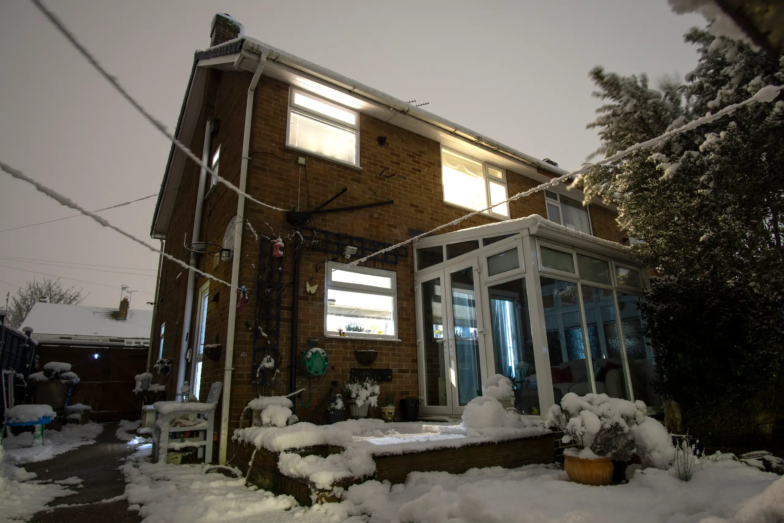 A house exterior covered in snow, featuring a glass extension and outdoor lighting at night